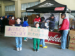 Two boys hold up a sign made from a cardboard box that reads, "Thank you, from all of us at the Utatsu Evacuation Center."